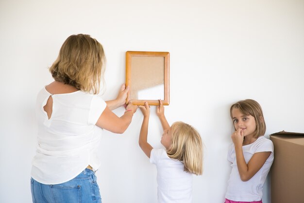 Encantadora niña poniendo marco en la pared blanca con la ayuda de mamá