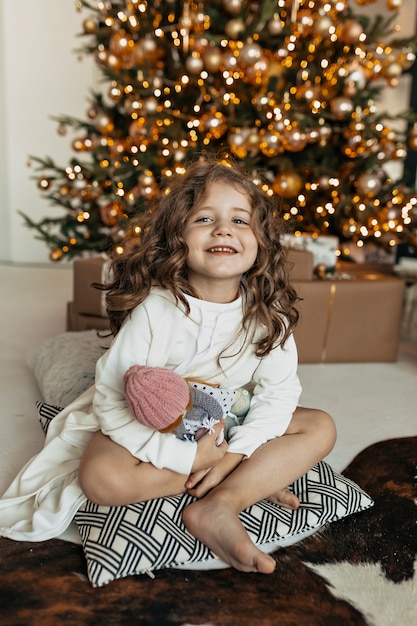Encantadora niña sentada en una almohada con juguete sobre el árbol de Navidad, humor de año nuevo, fiesta de Navidad