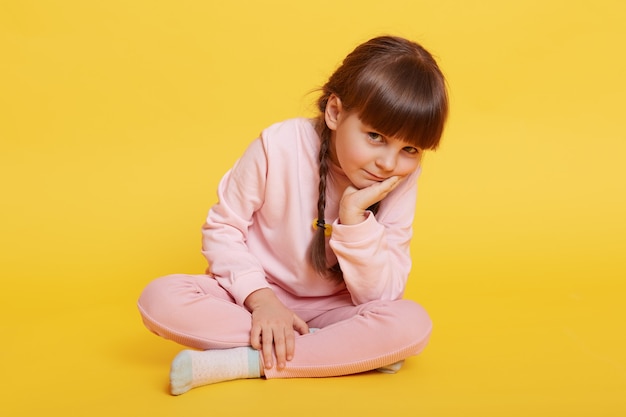 Encantadora niña sentada en el piso con las piernas cruzadas, manteniendo la palma en la barbilla, mirando aburrida a la cámara, la niña vestida con ropa casual rosa, el niño quiere jugar, no sabe qué hacer.