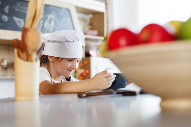 Encantadora niña en un sombrero de jefe se encuentra en una mesa lista para cocinar