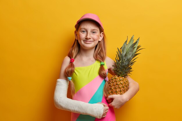 Encantadora niña en traje de baño de colores y gorra, posa con piña contra la pared amarilla, disfruta del verano y buen descanso, tiene un brazo roto después de caer desde una altura o un deporte peligroso