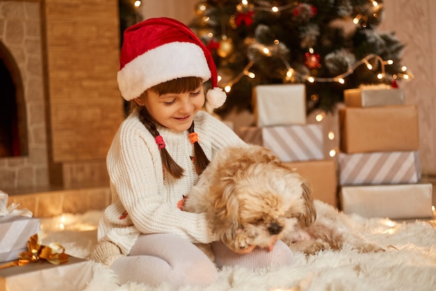 Encantadora niña vestida con suéter blanco y sombrero de santa claus, jugando con su cachorro mientras está sentada en el piso cerca del árbol de Navidad, cajas de regalo y chimenea.