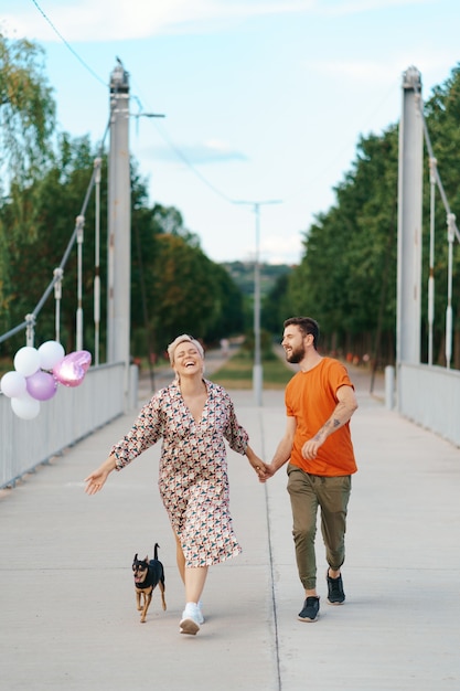 Encantadora pareja alegre caminando feliz en el puente con su perro y globos rosados sonriendo