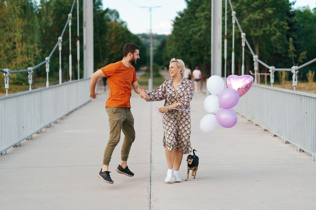 Encantadora pareja alegre caminando feliz en el puente con su perro y globos rosados sonriendo