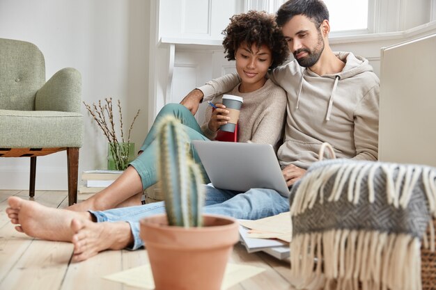 Encantadora pareja familiar se abrazan, se visten de manera informal, disfrutan del ambiente doméstico, sincronizan datos en la computadora portátil, trabajan en proyectos de negocios familiares, beben bebidas calientes, cactus en primer plano