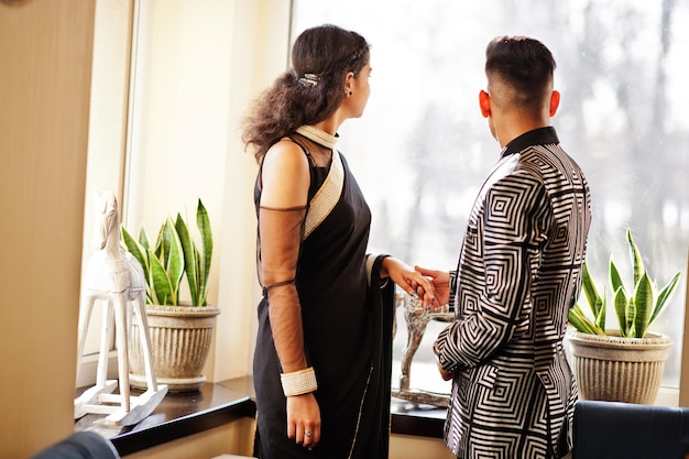 Foto gratuita encantadora pareja india enamorada vestida con sari y elegante traje posado en el restaurante detrás de ellos miran por la ventana y se toman de la mano