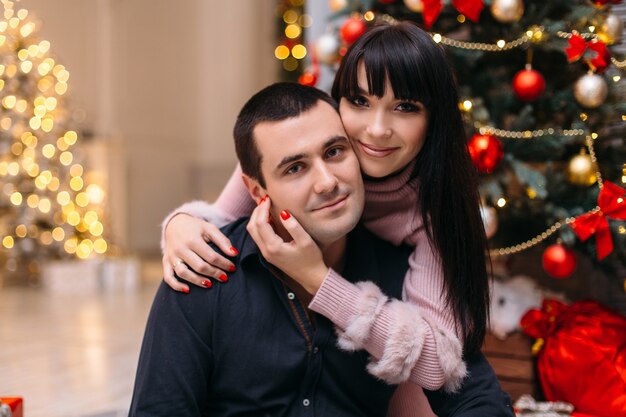 Encantadora pareja joven feliz posa ante un árbol de Navidad rojo en un rincón acogedor