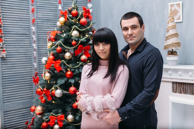 Encantadora pareja joven feliz posa ante un árbol de Navidad rojo en un rincón acogedor