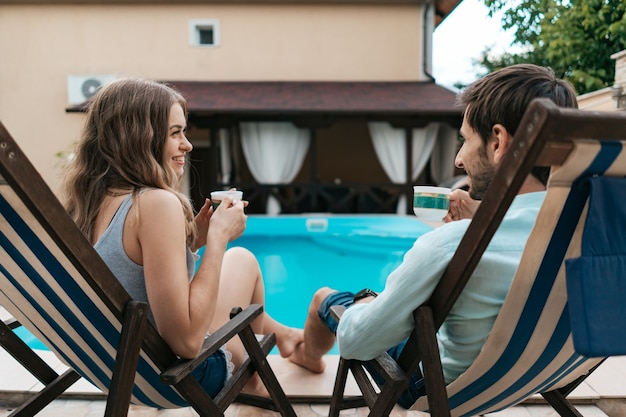 Encantadora pareja joven pasar tiempo juntos en casa cerca de la piscina mientras charla y disfruta del té