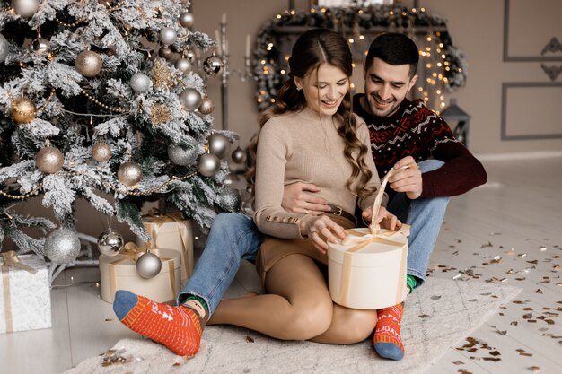 Encantadora pareja joven en ropa de hogar acogedor abre cajas presentes antes de un árbol de Navidad