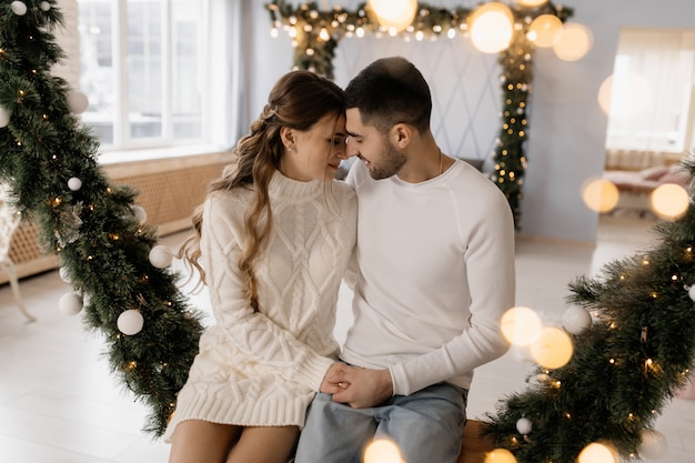 Encantadora pareja joven en ropa de hogar blanca acogedora posa en una habitación con árbol de Navidad