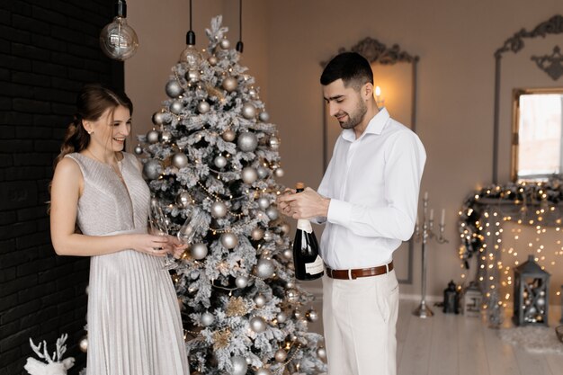 Encantadora pareja joven en ropa de lujo posa con copas de champán ante un árbol de Navidad