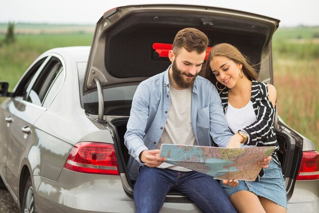 Encantadora pareja de jóvenes sentados en el tronco del coche buscando destinos en el mapa