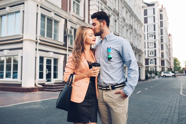 Encantadora pareja paseando por el barrio británico. Hombre de cabello oscuro con camisa azul besos en la cabeza chica rubia vestida de negro con chaqueta coral.