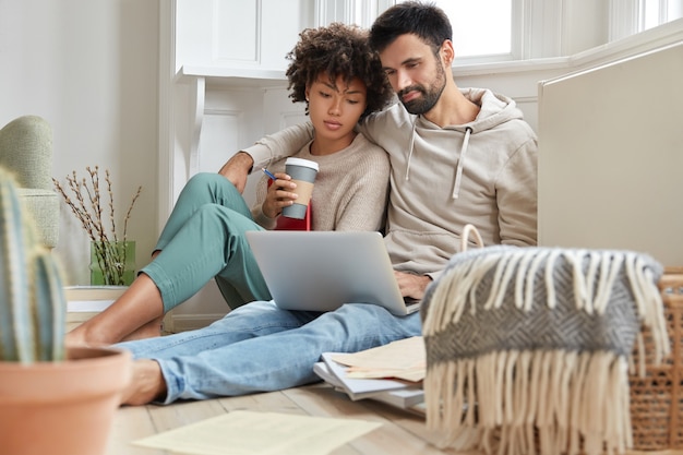 Encantadora pareja de raza mixta se abrazan, se sientan en el piso, se sienten relajados mientras ven una película en la computadora portátil