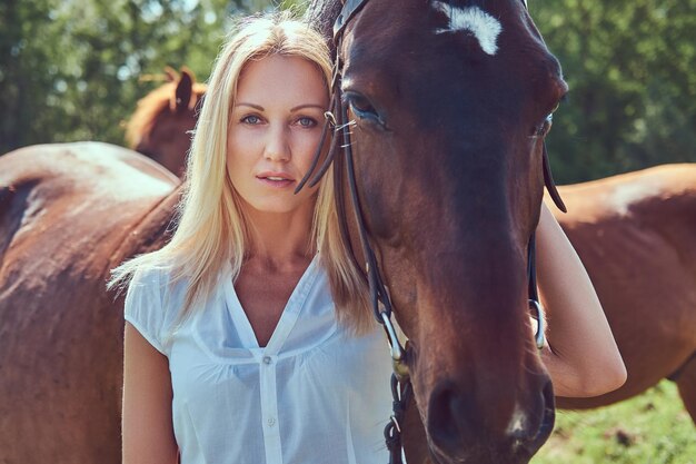 Encantadora rubia hermosa con una blusa blanca y jeans abrazando un caballo a un campo verde.