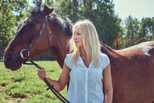 Encantadora rubia hermosa con una blusa blanca y jeans de pie con un caballo en un campo.