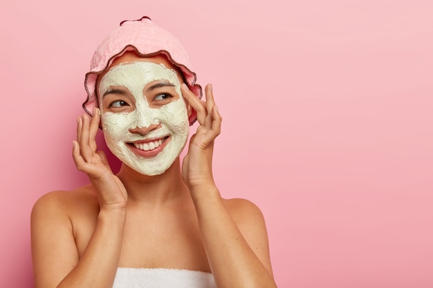 Encantadora y satisfecha modelo femenina asiática usa mascarilla facial para una piel suave y perfecta, posa con gorro de ducha y toalla después del baño, toca las mejillas, mira con alegría a un lado. Concepto de bienestar y perfección