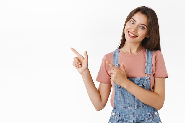 Encantadora, seductora, alegre y hermosa mujer con camiseta de peto inclinando la cabeza tonta y sonriendo mientras promociona el producto de la empresa apuntando a la esquina superior izquierda da consejos para ver un anuncio genial