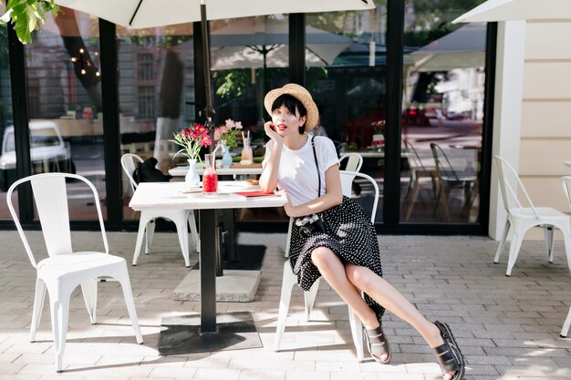 Encantadora señorita con sombrero de verano descansando en un café al aire libre con la cara apoyada con la mano y esperando amigo