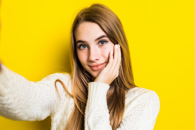 Encantadora sonriente chica de moda soleada está haciendo selfie en su teléfono con un suéter blanco ancho en amarillo