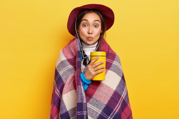 La encantadora y sorprendida viajera tiene un descanso activo para acampar, se calienta con una bebida caliente y una tela escocesa a cuadros, mantiene los labios doblados