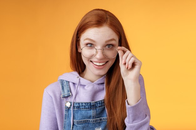 Encantadora tonta modesta joven pelirroja friki amante de los juegos discus últimas tendencias de juego sonriendo felizmente divertidas gafas táctiles sonriendo curiosamente reciben lindo regalo sorprendido, fondo naranja