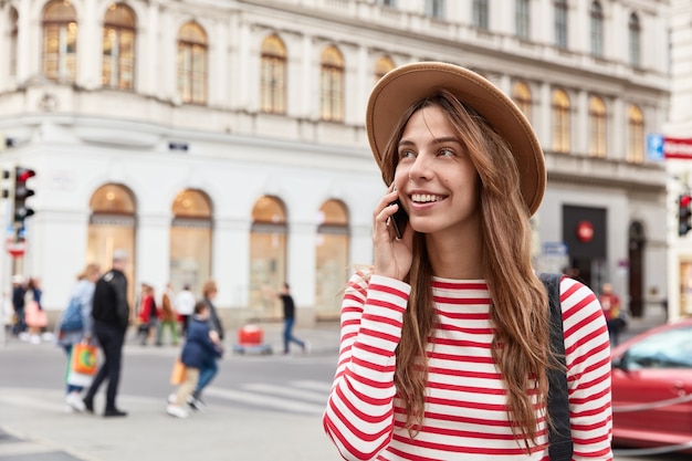 Encantadora viajera camina en un entorno urbano, llama a un amigo en un dispositivo smartphone moderno