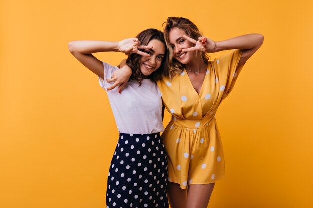 Encantadoras damas de buen humor abrazándose en amarillo. Amigas con estilo posando con el signo de la paz y riendo.