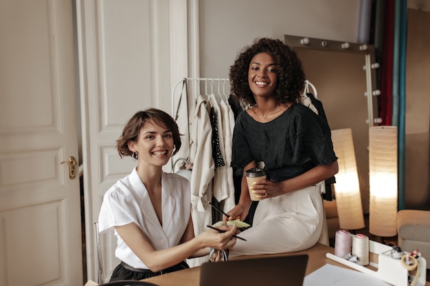 Encantadoras damas vestidas en blanco y negro sonríen y trabajan en un nuevo diseño de ropa