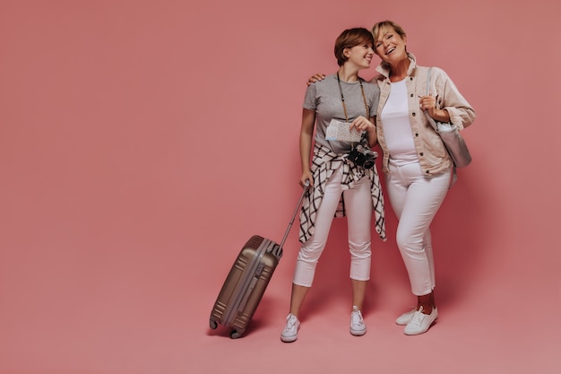 Encantadoras dos damas con peinado corto y fresco en ropa ligera y moderna posando con boletos, cámara y maleta y sonriendo sobre fondo rosa.