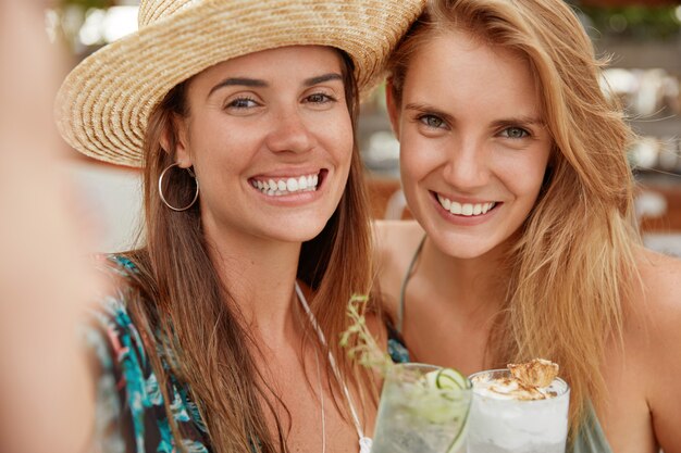 Encantadoras hembras alegres con amplias sonrisas, posan para selfie, beben cócteles refrescantes fríos. Las blogueras relajadas se recrean en un país tropical. Las mujeres jóvenes positivas se hacen fotos