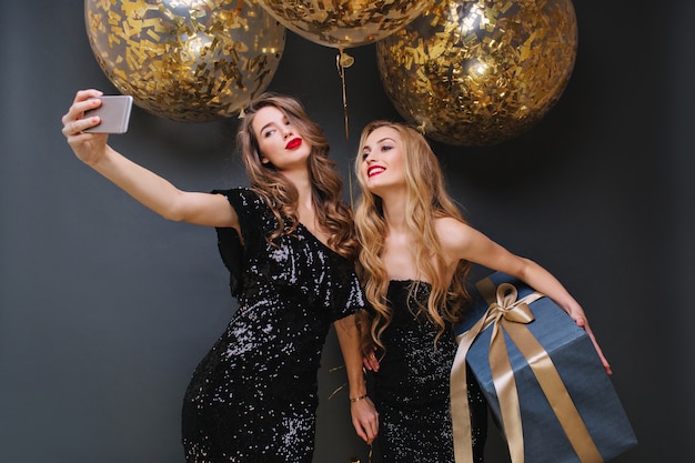 Encantadoras mujeres jóvenes de moda en vestidos negros de lujo haciendo selfie con globos grandes con oropeles dorados. Divertirse, presentar, expresar positividad, sonreír.