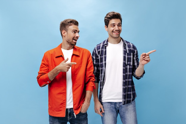 Encantadores hombres barbudos alegres apuntan al lugar para el texto en la pared azul. Retrato de chico con chaqueta naranja y su amigo en camisa a cuadros.