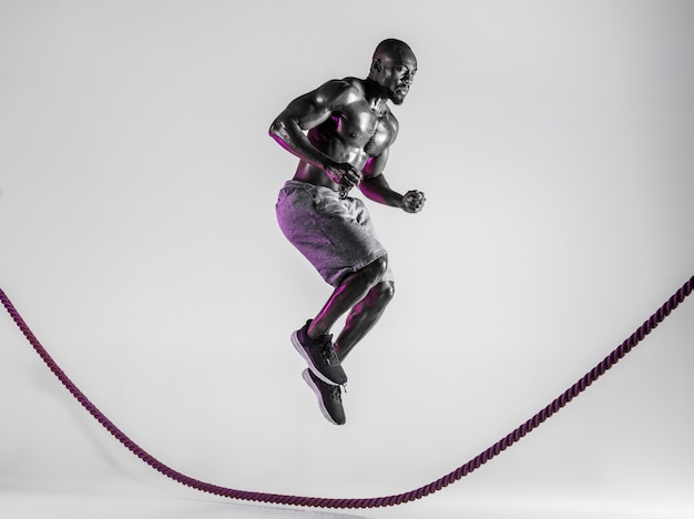 Por encima del mundo. Entrenamiento joven culturista afroamericano sobre fondo gris de estudio. Modelo masculino musculoso en ropa deportiva saltando por encima de la cuerda de batalla. Concepto de deporte, culturismo, estilo de vida saludable.