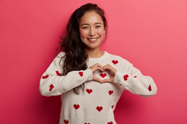 Encontraste la llave de mi corazón. Hermosa mujer coreana sonriente hace un signo de amor, expresa sentimientos tiernos, tiene cola de caballo, piel sana, usa un suéter, tiene un estado de ánimo romántico. Mujeres