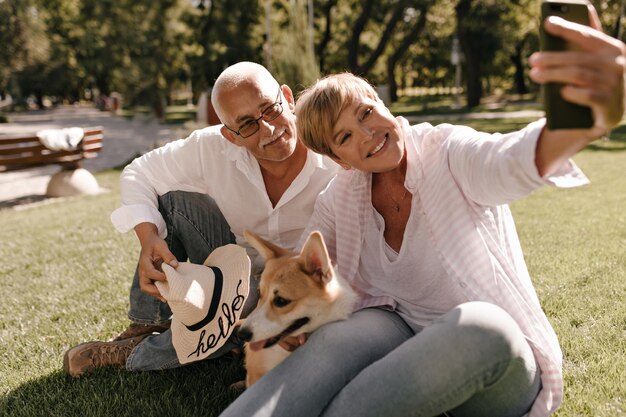 Endecha moderna con un peinado corto y fresco en una blusa rosa sonriendo sentada en el césped y tomando una foto con un hombre con anteojos y un perro en el parque