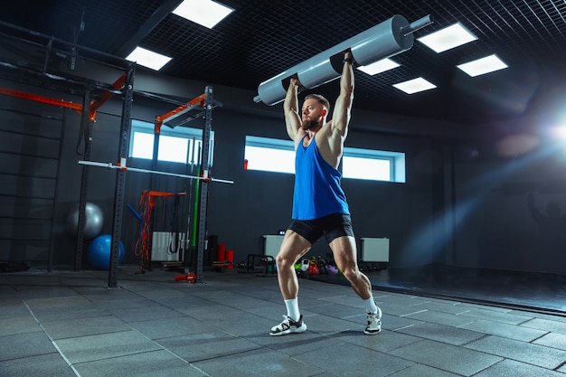 Energía salvaje. Joven atleta caucásico muscular entrenando en el gimnasio, haciendo ejercicios de fuerza, practicando, trabajando en la parte superior de su cuerpo con pesas y barras. Fitness, bienestar, concepto de estilo de vida saludable.