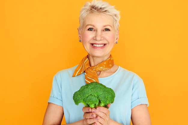 Enérgica hermosa mujer de mediana edad con pelo gris corto posando aislada con brócoli verde en sus manos, que va a hacer una ensalada orgánica saludable.