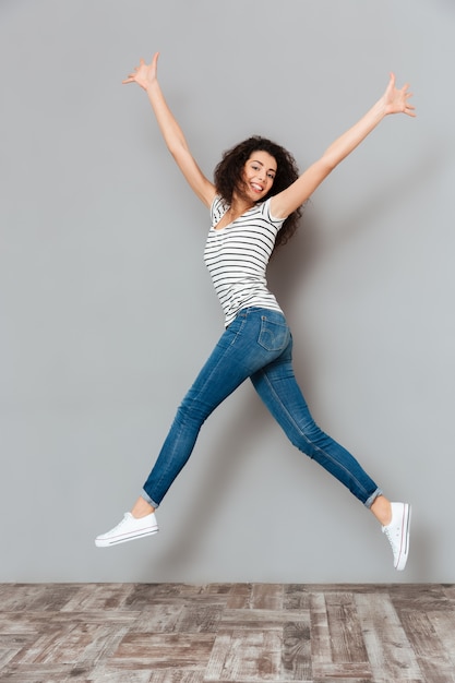 Enérgica mujer de 20 años en camiseta a rayas y jeans saltando con las manos vomitando en el aire sobre gris