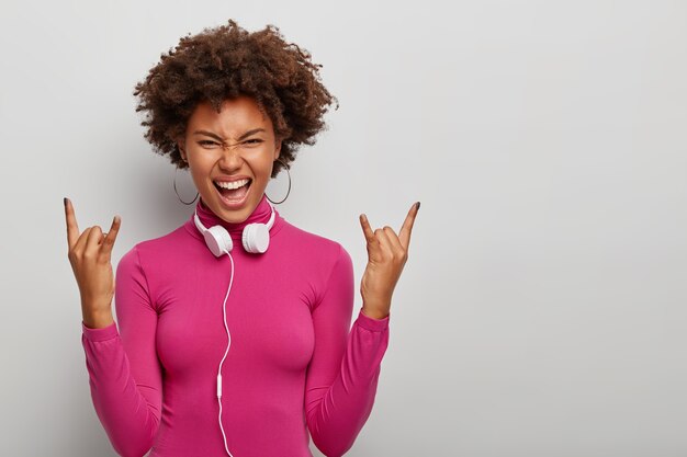 La enérgica mujer rizada meloman hace un gesto de rock n roll, tiene una alegre expresión rebelde