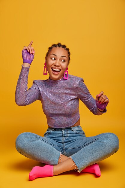 Enérgica y positiva chica de piel oscura de buen humor se sienta con las piernas cruzadas, levanta los brazos y se mueve con el ritmo de la música, mira felizmente a un lado, viste jersey, jeans y calcetines, aislado sobre una pared amarilla