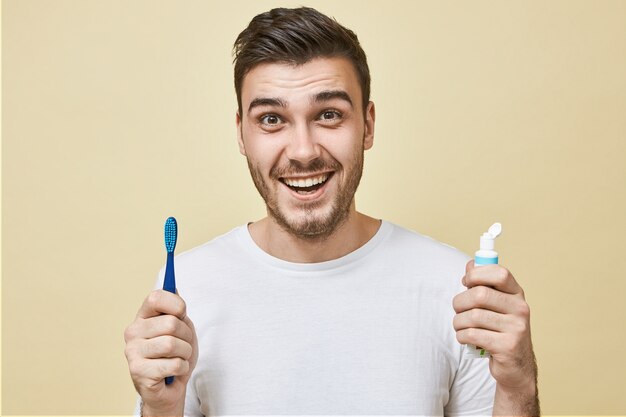 Enérgico joven positivo con rastrojo posando con cepillo de dientes y pasta blanqueadora sonriendo ampliamente con dientes blancos perfectos. Hábitos saludables, rutina diaria y cuidado dental