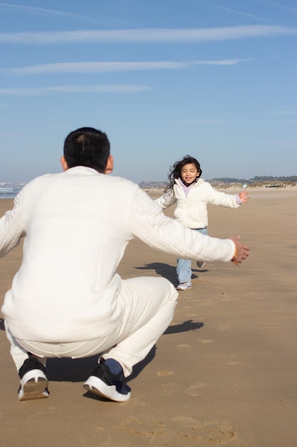 Enérgico padre e hija jugando en la playa. Familia japonesa en un día soleado, niña corriendo hacia su padre. Papá ocio, tiempo en familia, concepto de crianza