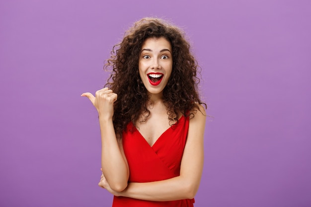 Energizada atractiva y asombrada mujer guapa con peinado rizado sonriendo asombrada, sorprendida apuntando a la izquierda con el pulgar preguntando acerca de la exhibición interesante en vestido rojo sobre la pared púrpura.