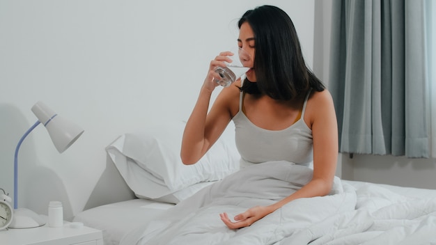 La enferma enferma india sufre de insomnio. Joven asiática tomando analgésicos para aliviar el dolor de cabeza y beber un vaso de agua sentado en la cama en su habitación en casa por la mañana.