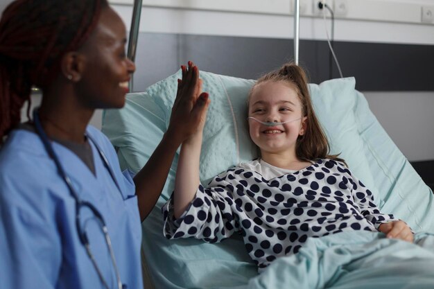 Enfermera afroamericana haciendo un gesto alto con una niña enferma descansando en la cama del paciente de la sala pediátrica del hospital. El personal del centro de atención médica para niños choca los cinco con un niño enfermo bajo tratamiento médico.