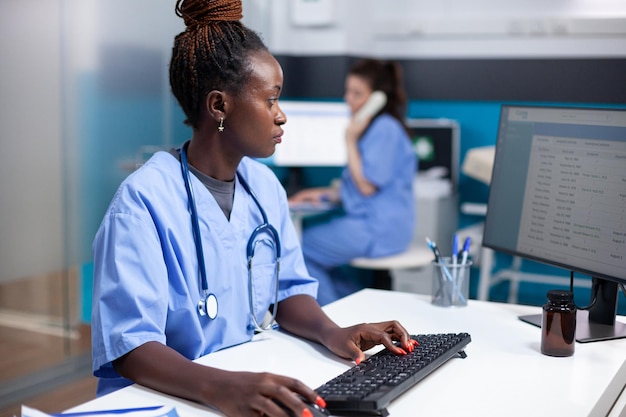 Foto gratuita enfermera afroamericana trabajando en el escritorio, insertando datos en la lista de citas. mujer adulta concentrada comprobando la lista de pacientes, usando estetoscopio en una oficina médica limpia y moderna que opera computadora