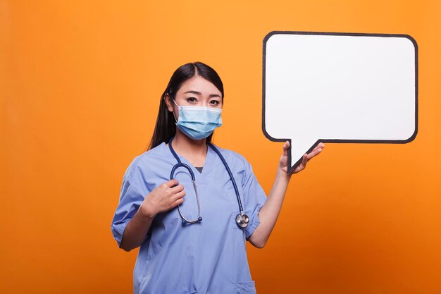 Enfermera asiática confiada de la clínica de salud para adultos jóvenes con mascarilla y uniforme azul mientras sostiene una burbuja de habla de cartón. Cuidador del hospital con cartel de burbuja de habla de cartón.