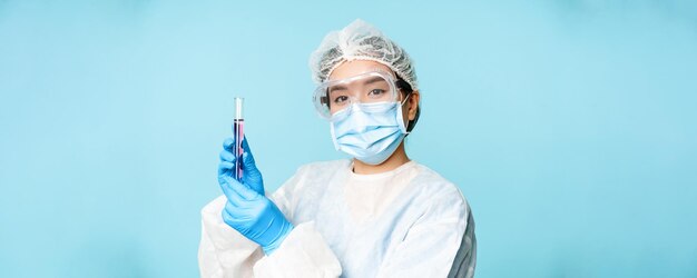 Enfermera asiática o trabajador de laboratorio en equipo de protección personal que muestra un tubo de muestra de prueba de pie en una máscara facial médica sobre fondo azul.